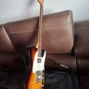 Elektrická kytara s tónem sunburst (černá, červená a žlutá) a bílým pickguardem. Na hlavě hředu je viditelné logo Fender. Kytara je umístěna před ote…