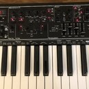 Černý syntetizátor s bílými a černými klávesami. Horní panel obsahuje ovládací prvky rozdělené do sekcí Oscillators, Mixer, Filters, Porta, LFOs a En…