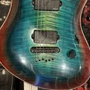 Elektrická kytara s modrozelým plamenovým javorovým topem a hnědými okraji. Nástroj má dva černé humbuckery, na kterých je viditelný nápis FISHMAN. K…