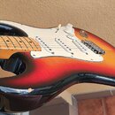 Elektrická kytara s tělem v červeno-žlutém gradientu (sunburst) a černým pickguardem. Na bílém plastovém krytu jsou tři snímače a ovládací prvky. Na…