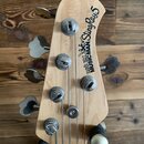 Hlava pětistrunné basové kytary z přírodního světlého dřeva. Na povrchu je černý nápis ERNIE BALL MUSIC MAN StingRay 5 doprovázený logem postavy. Jso…