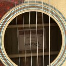 Detail znazorněný skrze otvor akustické kytary. Uvnitř je vidět štítek s nápisy Takamine a GD50-NAT. Kytara má světle dřevěnou barvu, vlnitý ornament…