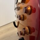 Detailní pohled na pravou stranu těla elektrické kytary v červeně gradientním lakování. Na povrchu jsou čtyři hnědé plastové ovládací knofíky s čísli…