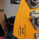 Záběr na zadní stranu hlavy kytary z jasného dřeva s viditelnou masivkou. Na dřevě je černý nápis: „Designed and Backed by Fender® s/n ISSE21003417 C…