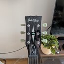 Černá hlava kytary s bílým nápisem Harley Benton. Na přední straně je umístěn bílý kosočtvercový ornament a v dolní části logo HB. Jsou vidět tři lad…