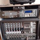 Fotografie zachycuje rack s audio vybavením, nad kterým je umístěn černý reprodukník značky BEHRINGER. V racku se nachází CD přehrávač s nápisem KAST…
