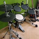 Elektronická bicí souprava s černými gumovými pady a činely na stříbrném kovovém rámu. Na bílém bubnu je viditelný nápis OXO. Na pedálu basového bubn…