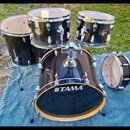 Sada tmavě modrých bubnů značky TAMA. Soubor obsahuje basový buben, dva tom-tomy, floor tom a malý buben. Na basovém bubnu je bílý nápis TAMA. Povrch…