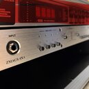 Stříbrný rackový zesilovač s nápisem ZMACS 4X4. Na předním panelu je vstup označený jako INPUT a čtyři malé ovládací knoflíky pod nápisem AMPLIFIER.…