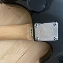 Detail zadní strany černé kytary s dřevěným hřádem. Na spojení krku a těla je nalepený zlatý štítek s nápisem MODEL BT-2BK. Vedle něj je kovový kryt…