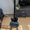 Černá elektrická kytara s dvojstupňovou hlavou a bílými ladičkami. Na hlavě je viditelné logo STEREO a nápis MODEL 100. Kytara stojí na stojanu v int…