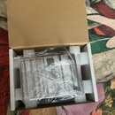 Otevřený hnědý papírový krabici s bílým polystyrenovým vložkem. Uvnitř je v průhledné plastové fólii zabalený předmět s doprovodnou dokumentací. Text…