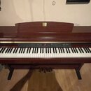 Digitální piano v tmavě hnědém dřevěném provedení s bílými a černými klávesami. Na horní části je umístěno zlaté logo s nápisem "Clavinova". Ovládací…