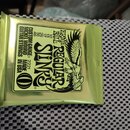 Zlatavě lesklé sáček s kytarovými strunami. Na obalu je černý nápis ERNIE BALL REGULAR SLINKY a CUSTOM GAUGE 2221 NICKEL WOUND GUITAR STRINGS MADE IN…