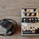 Kytarový efekt v bílém provedení s černým panelem a připojeným externím zdrojem napájení. Na černém panelu je bílý nápis „Wiggler“ a text „electro-ha…