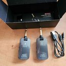 Dva šedé bezdrátové vysílače a přijímače s kovovými konektory, na kterých je nápis NUX B-2 PLUS. Vedle nich je černý USB kabel a v pozadí otevřená če…