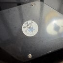 Černý kovový nebo plastový kryt s rounded hranami, připevněný čtyřmi šrouby. Uprostřed je nalepená kulatá bílá nálepka s nápisem „Inspected By“ a ruč…