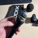 Ruka drží černý mikrofon bez mřížky, na kterém je bílá nálepka s nápisy AUDIX a MADE IN USA. V pozadí je černý obal s bílým nápisem AUDIX, samostatná…