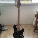 Elektrická basová kytara s černým tělem a světlou dřevěnou hmatníkem. Na hlavě je viditelné logo Fender. Nástroj stojí na černém stojanu v interiéru.
