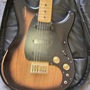 Elektrická kytara s tónem sunburst a černým pickguardem, položená v černém pouzdře. Nástroj má světle hnědý javorový hmatník a kovový most. Na kytaru…