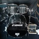 Černá bicí souprava značky SONOR, model SQ2. Sestava obsahuje basový buben s nápisem SONOR a SQ2, dva tom-tomy a jeden floor tom. Vedle nástrojů jsou…