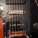 Detail elektrické kytary s černým pickguardem a hnědým tělem. Jsou vidět dva černý humbuckery, chromovaný kobylník a jeden černý ovládací knoflík s n…
