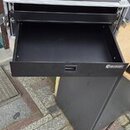 Černý transportní kufr s hliníkovými hranami a kovovými rohy. Kufr je otevřený a má vysunutou spodní zásuvku. Na pravém boku zásuvky je viditelné bíl…