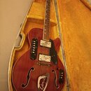 Polodupatá elektrická kytara v červeném provedení s f-otvory, položená v otevřeném kufru s žlutým vnitřkem. Na těle je černý pickguard a kovový ozdob…