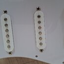 Bílá plechtráčka s třemi bílými snímači typu single-coil. Snímače jsou připevněny šrouby a mají kovové póly. Na fotce nejsou žádné viditelné značky a…