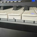 Detailní záběr na klávesy elektronického klavíru v černé barvě. Bílé plastové klávesy vykazují známky opotřebení, konkrétně jsou vidět stopy po škráb…