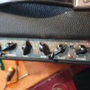 Detail ovládacího panelu zesilovače s kovovým povrchem a černými plastovými knoflíky. Jsou zde viditelné nápisy INPUT, VOLUME, TREBLE, BASS, MASTER a…