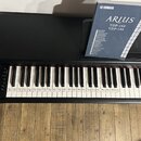 Černý digitální piano s bílými a černými klávesami. Na pravé straně je nápis YAMAHA, na přiloženém manuálu jsou viditelné texty YAMAHA, ARIUS a model…