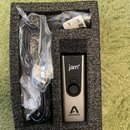 Černý obdélníkový přístroj značky APOGEE s nápisem jam+ umístěný v černé pěnové výplni. Zařízení má na přední straně ovládací knoflík a tlačítko. Ved…