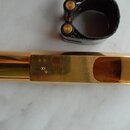 Zlatý kovový mouthpiece s otvoru pro rty. Na povrchu je vyryté symbol nekonečna, hvězdička a pod nimi nečitelný nápis. Vedle nástroje je umístěno čer…
