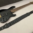 Černá elektrická basová kytara s hnědým hmatníkem a zlatým hardwarem. Na hlavě nástroje je viditelné logo Fender. Vedle kytary leží černý popruh s bí…
