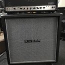 Kytarový zesilovač a reprobox. Na horním zesilovači je logo Peavey a nápis 6505. Spodní reprobox má šedou síťovku s bílým rámem a uprostřed nápis RIV…