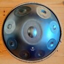 Kulatý kovový handpan s modravým, gradientním povrchem a několika vyhlubecinami (tóny). Nástroj má pletený lem v béžové barvě. V dolní části je bílý…