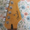 Fotografie zachicuje hlavu elektrické kytary s přírodním dřevěným povrchem a hnědým hmatníkem. Na hlavě je čitelné logo Fender® a nápis STRATOCASTER®…