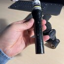 Černý mikrofon s kovovou mřížkou v držáku. Na horní části je bílá nálepka s nápisy AUDIO a MADE IN. Povrch černého těla vykazuje výrazné opotřebení v…