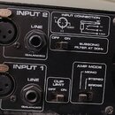 Zadávací panel černého zesilovače s dvěma vstupy. U obou vstupů jsou popisky INPUT 1 a INPUT 2, konektory XLR s nápisem PUSH a jackové vstupy označen…