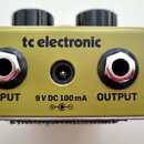 Záběr na zadní stranu olivově zeleného kytarového efektu. Na šasi je nápis tc electronic a tři konektory označené jako INPUT, OUTPUT a 9 V DC 100 mA…