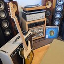 Na fotografii je elektrická kytara typu Telecaster s černým tělem a světlou hmatníkem, opřená o kytarový zesilovač s béžovým potahem. V pozadí jsou v…