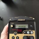 Černý spouštěcí modul s dorém panelu. Na horní straně je bílý nápis Roland a označení konektorů: OUTPUT L/MONO, R, TRIG IN 1, TRIG IN 2 a DC IN PSD A…