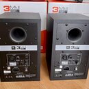 Dva černé studiové monitory v dřevěných skříních s kruhovým odstupňováním bassreflexu. Na zadní straně je bílý štítek s nápisem JBL 3 MkII POWERED ST…