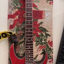 Elektrická kytara s červeným tělem a hnědým hmatníkem. Na těle je viditelný jeden černý humbucker. Kytara leží na tkaném textilu s vánočním motivem a…
