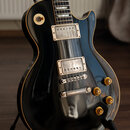 Černá elektrická kytara typu Les Paul s žlutým lemováním hran. Nástroj má dva humbuckery s krémovými rámečky, čtyři zlaté otočné knoflíky a stopku. P…