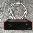 Červené audio rozhraní značky Focusrite s černým zadním panelem. Jsou zde viditelné konektory Input 2, Outputs L a R oraz USB port. Vedle zařízení le…
