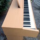 Digitální piano v dřevěné provedení světle hnědé barvy. Nástroj má standardní klaviaturu s černými a bílými klávesy a přiložený notový pult. Na vidit…