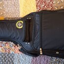 Černý pol pevný pouzdro na kytaru s žlutými ozdobnými lemovými pruhy. Na přední straně je kulatý štítek s nápisem „RockCase“. Pouzdro má několik zipo…
