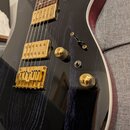 Elektrická kytara s černým lakem, pod kterým je viditelná struktura dřeva. Nástroj má zlatý hardware, včetně dvou humbuckerů a mostu. Na mostu je vid…
